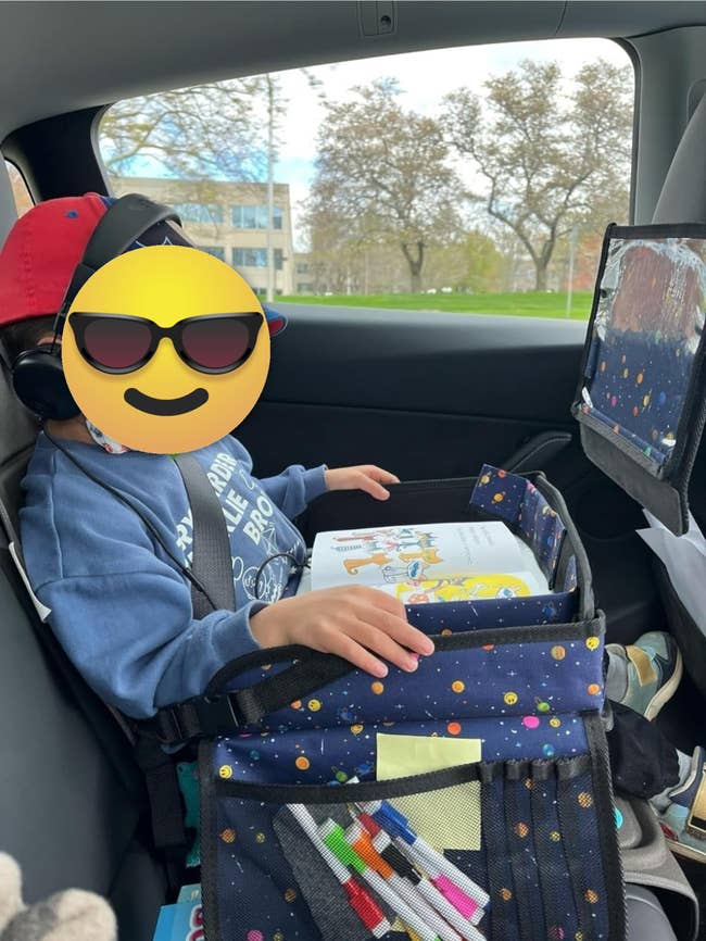 A young kid sits successful a car wearing headphones and a cap, utilizing a question enactment tray with drafting supplies