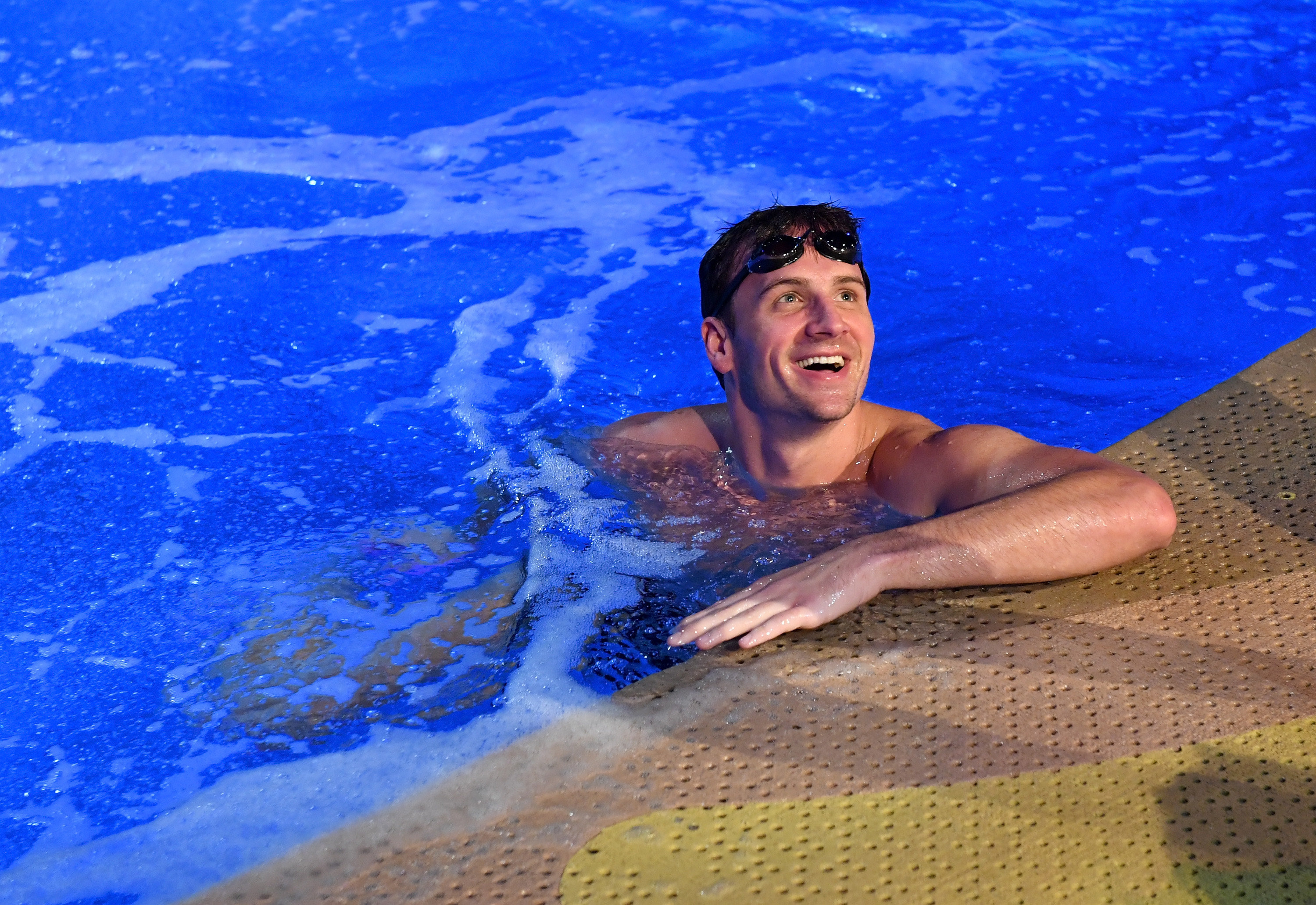 Ryan Lochte is in a swimming pool, resting his arms on the edge and smiling. He has goggles on his forehead. There is blue water around him