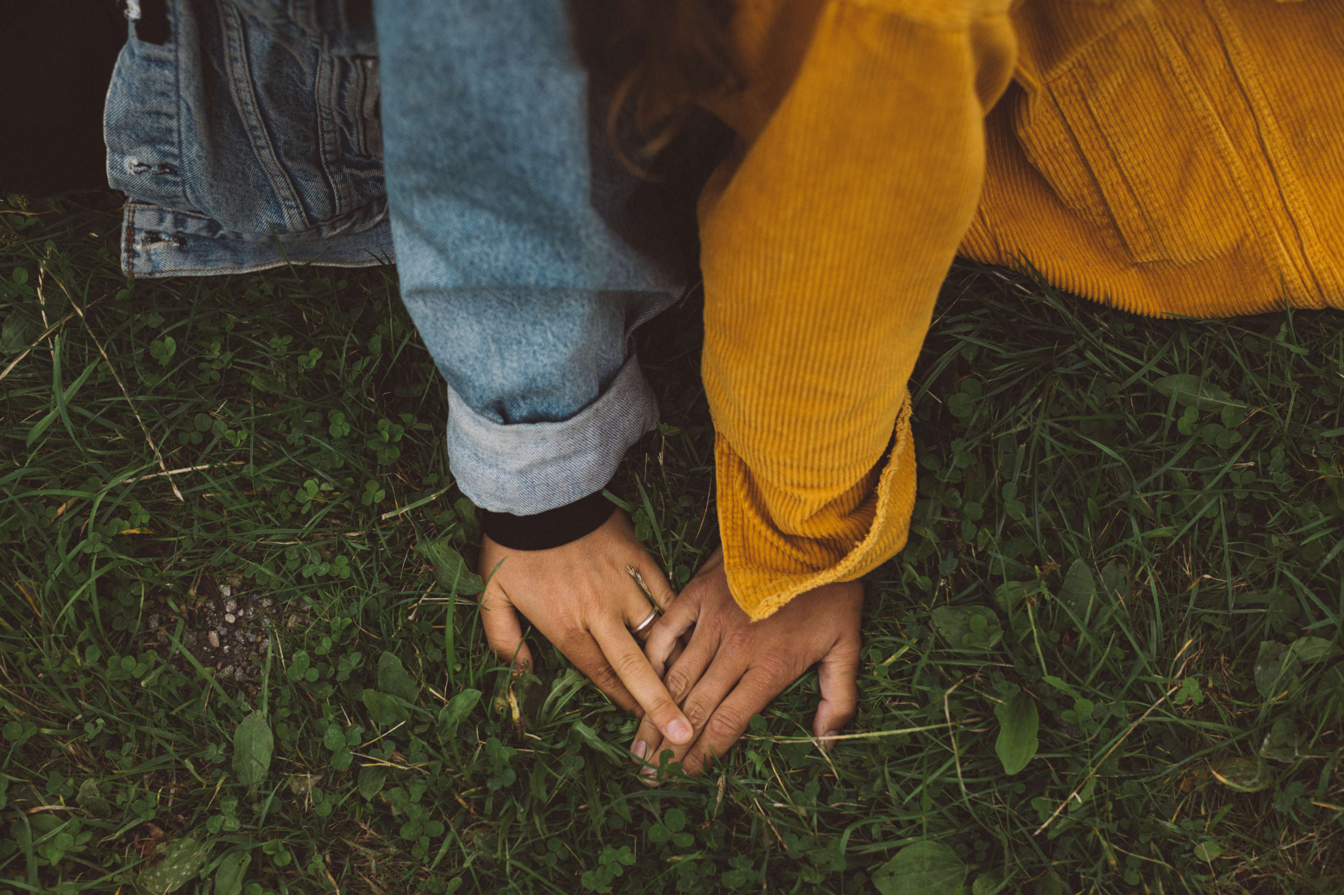 Two people laying on grass holding hands; one wears a denim jacket, the other a mustard-yellow sweater. Only their hands and part of their arms are visible