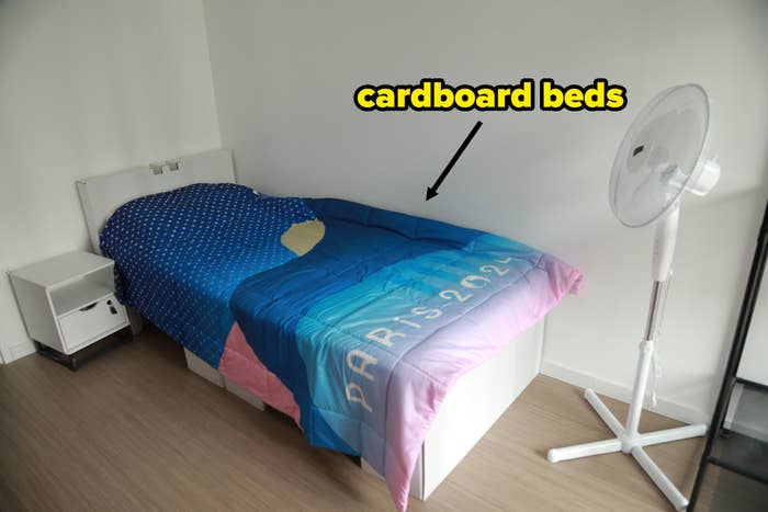 A simple bedroom with a single bed featuring a comforter that has &quot;Paris 2024&quot; written on it, a bedside table, and a fan