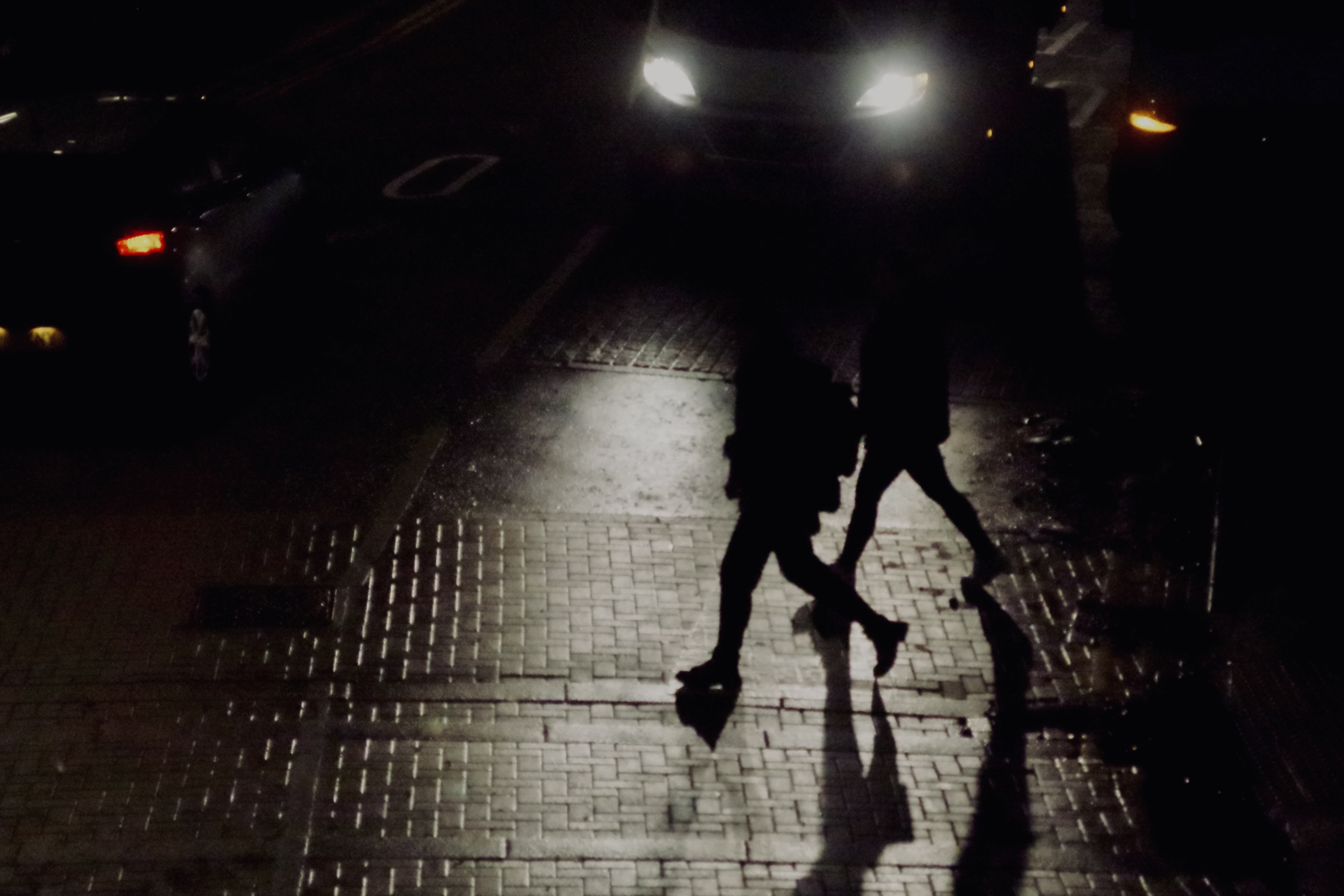 Two people are walking on a dimly lit street at night with car headlights in the background. Their identities are not discernible