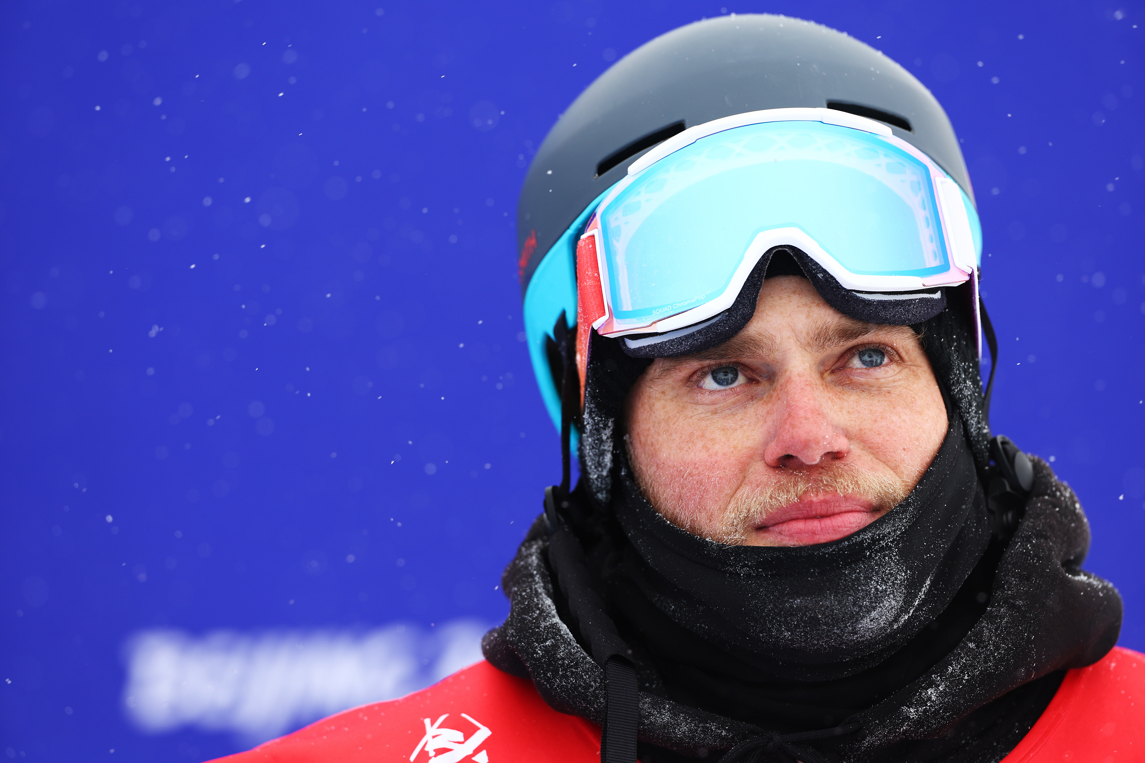 A person wearing ski goggles, a helmet, and a winter jacket, standing in front of a snowy background