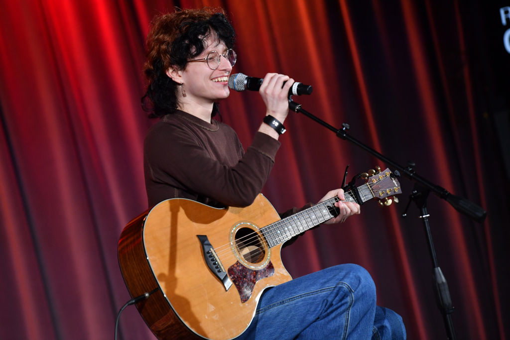 Cavetown playing an acoustic guitar while seated, smiling and singing into a microphone on stage. They are wearing glasses, a long-sleeved shirt, and jeans