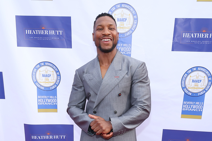 Jonathan Majors smiles on the red carpet in a gray double-breasted suit and black shoes at an event with NAACP and Beverly Hills Hollywood NAACP logos