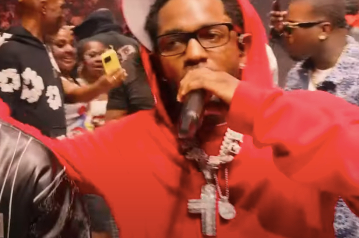 Kendrick Lamar (right) in a red hoodie, wearing glasses and holding a microphone