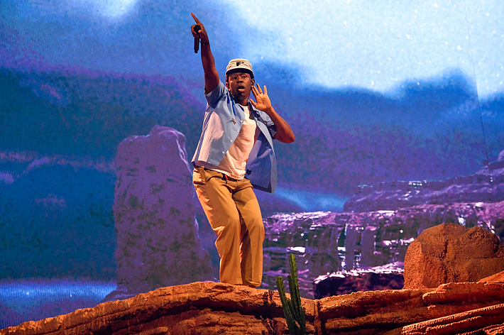 Tyler, the Creator performs on stage, wearing a blue hat, blue shirt, and beige pants, with a backdrop resembling a desert landscape