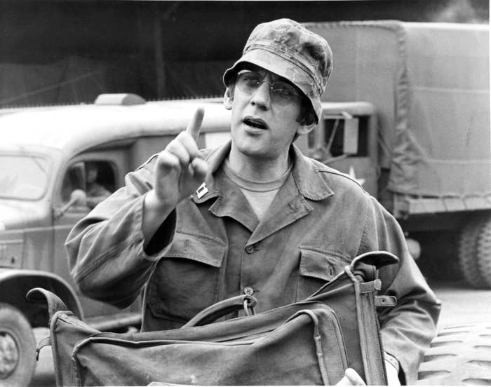 A man in military uniform and cap gestures with his finger and holds a bag, standing in front of military transport vehicles