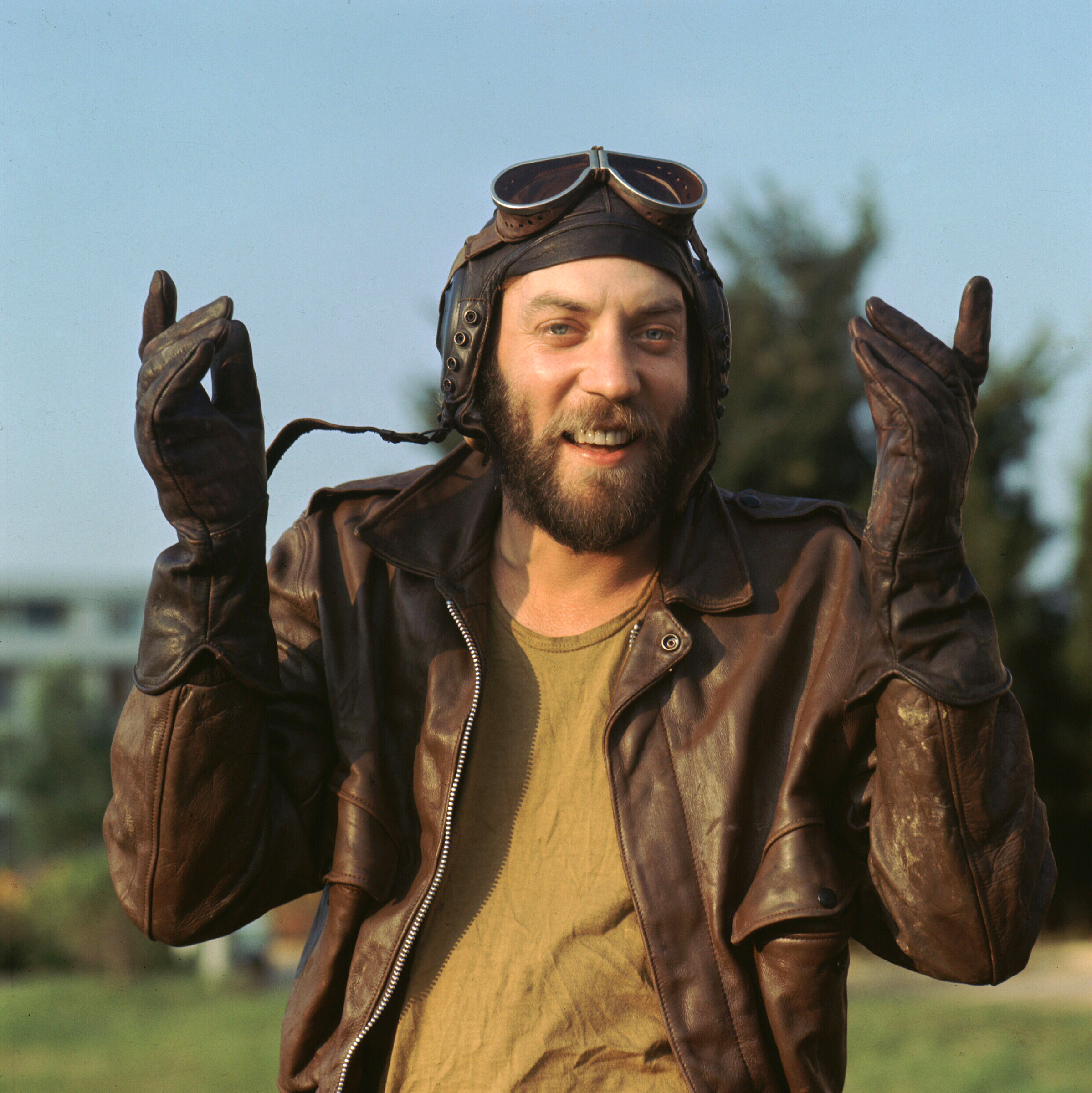 A bearded man in a leather pilot jacket and gloves smiles while gesturing with both hands in an outdoor setting, wearing aviator goggles on his head