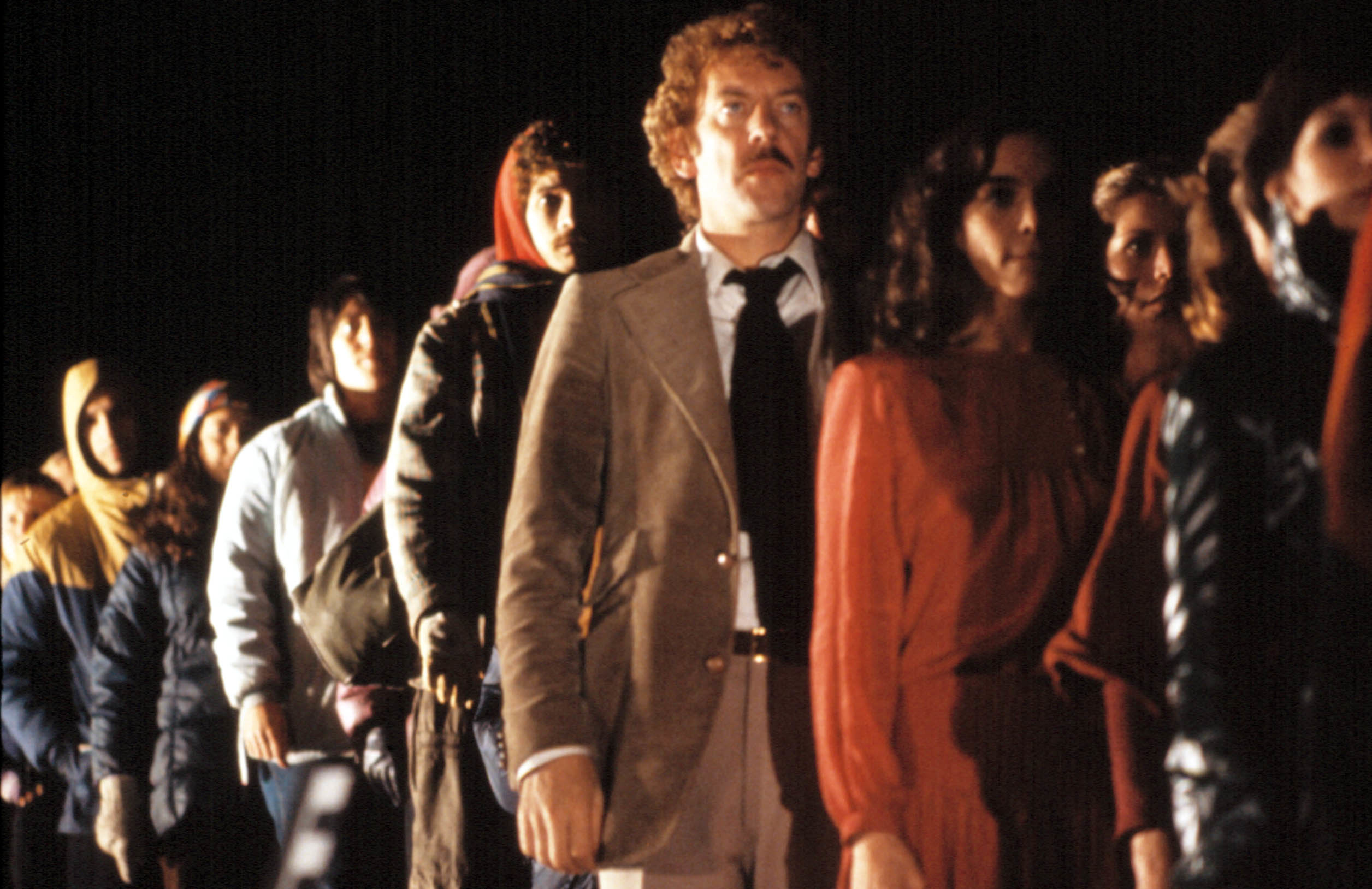 A line of people, including Donald Sutherland and Brooke Adams, stands in a dimly lit scene from the film "Invasion of the Body Snatchers."