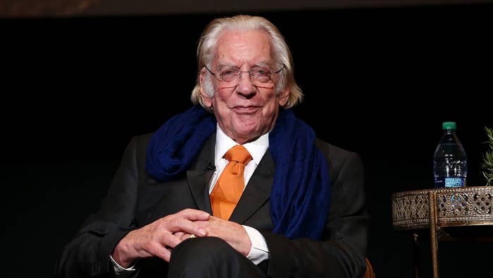 Donald Sutherland seated, wearing a dark suit, orange tie, and blue scarf, smiling at an event. A table with a water bottle is beside him