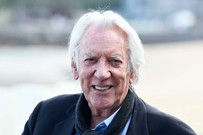 Donald Sutherland is smiling while wearing a dark jacket and a scarf. He appears outdoors