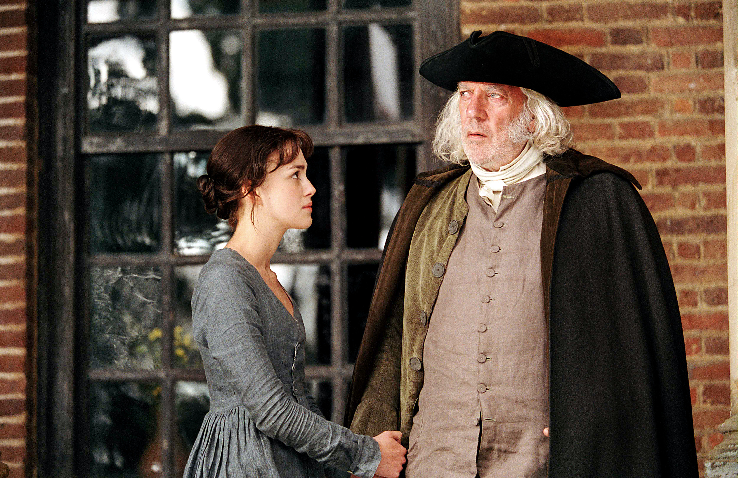 Keira Knightley holding hands with Donald Sutherland in a period costume scene, gazing at each other with emotion outside a brick building