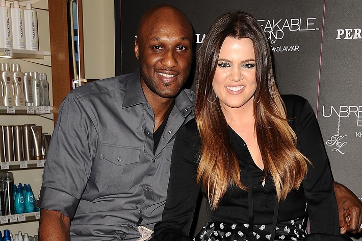 Lamar Odom and Khloé Kardashian pose together in front of perfume displays. Khloé wears a black blouse and patterned skirt