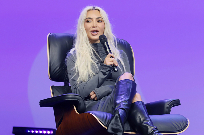 Kim Kardashian sits on a modern chair in a stylish outfit, holding a microphone, speaking at an event