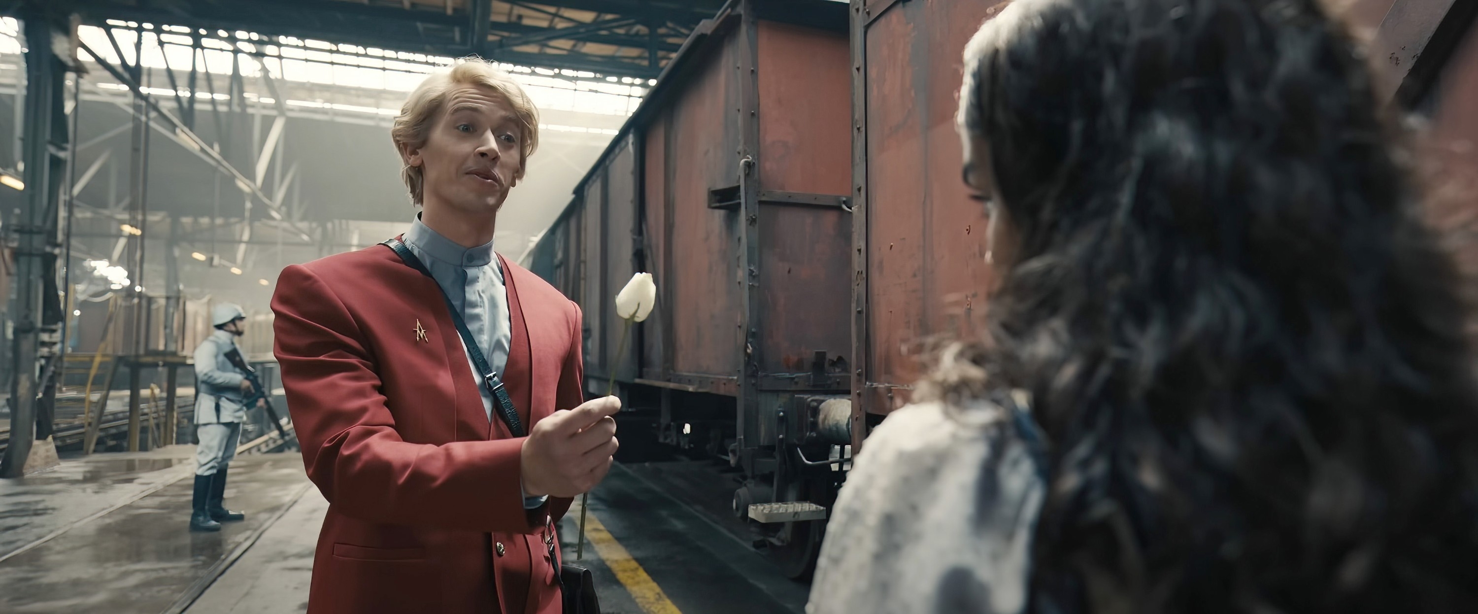 Tom Blyth, in a formal red outfit, holding a flower while interacting with a woman inside an industrial setting in a scene from the film "The Ballad of Songbirds &amp; Snakes"