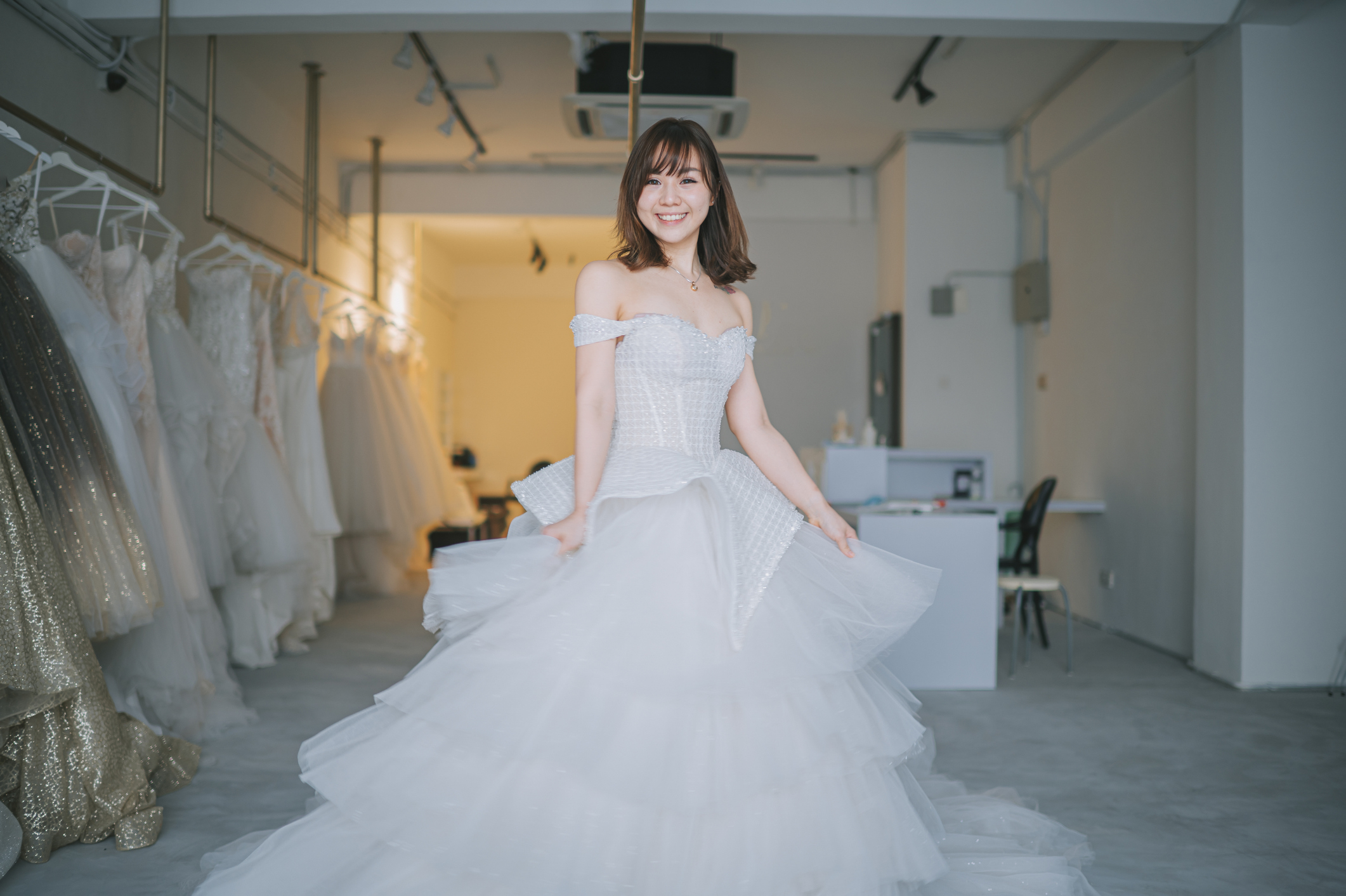 A bride in an off-shoulder, voluminous wedding gown smiles while posing in a bridal shop surrounded by wedding dresses