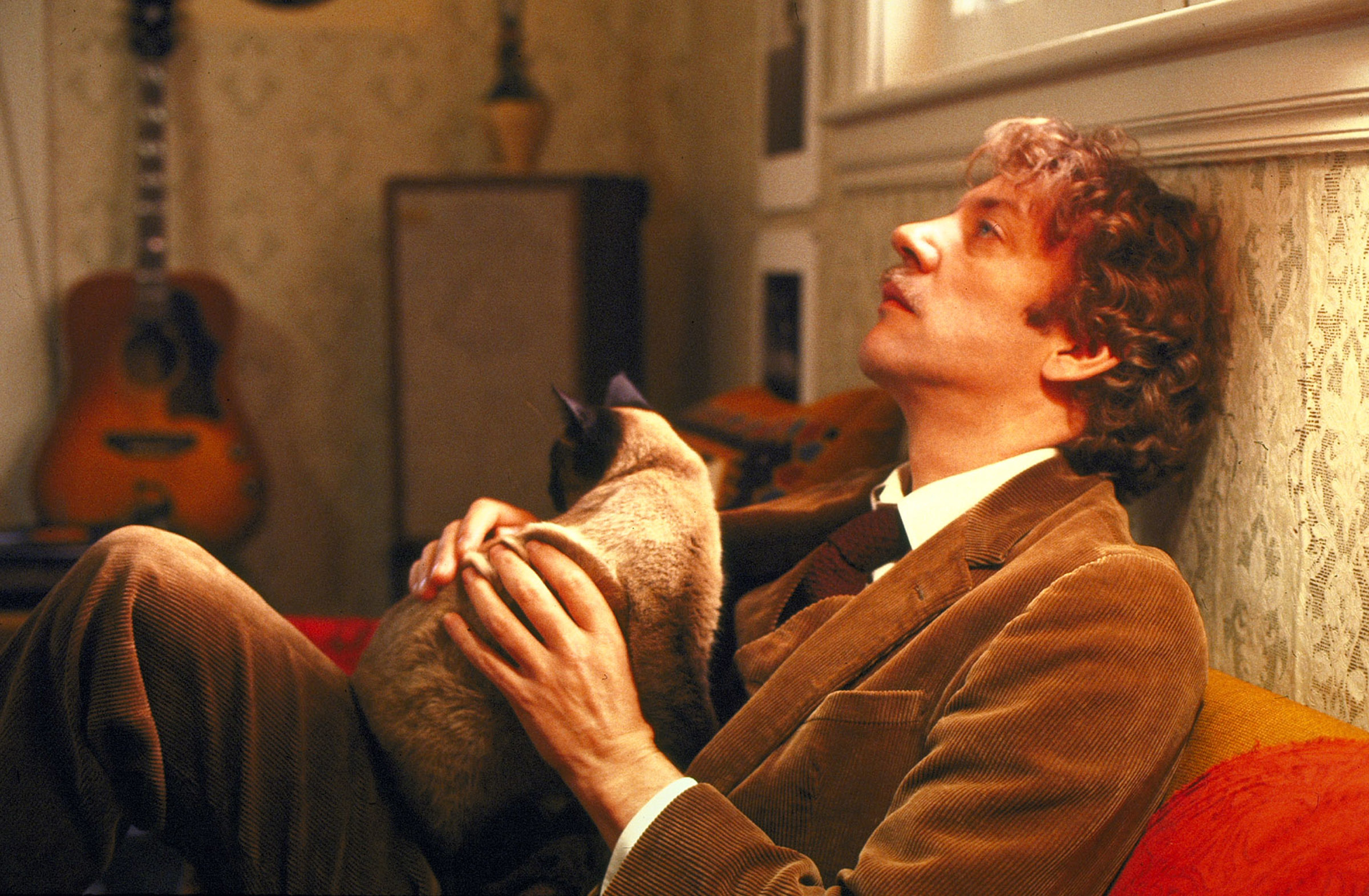 Man in corduroy suit sitting with Siamese cat on lap, looking contemplative in a cozy room with a guitar in the background
