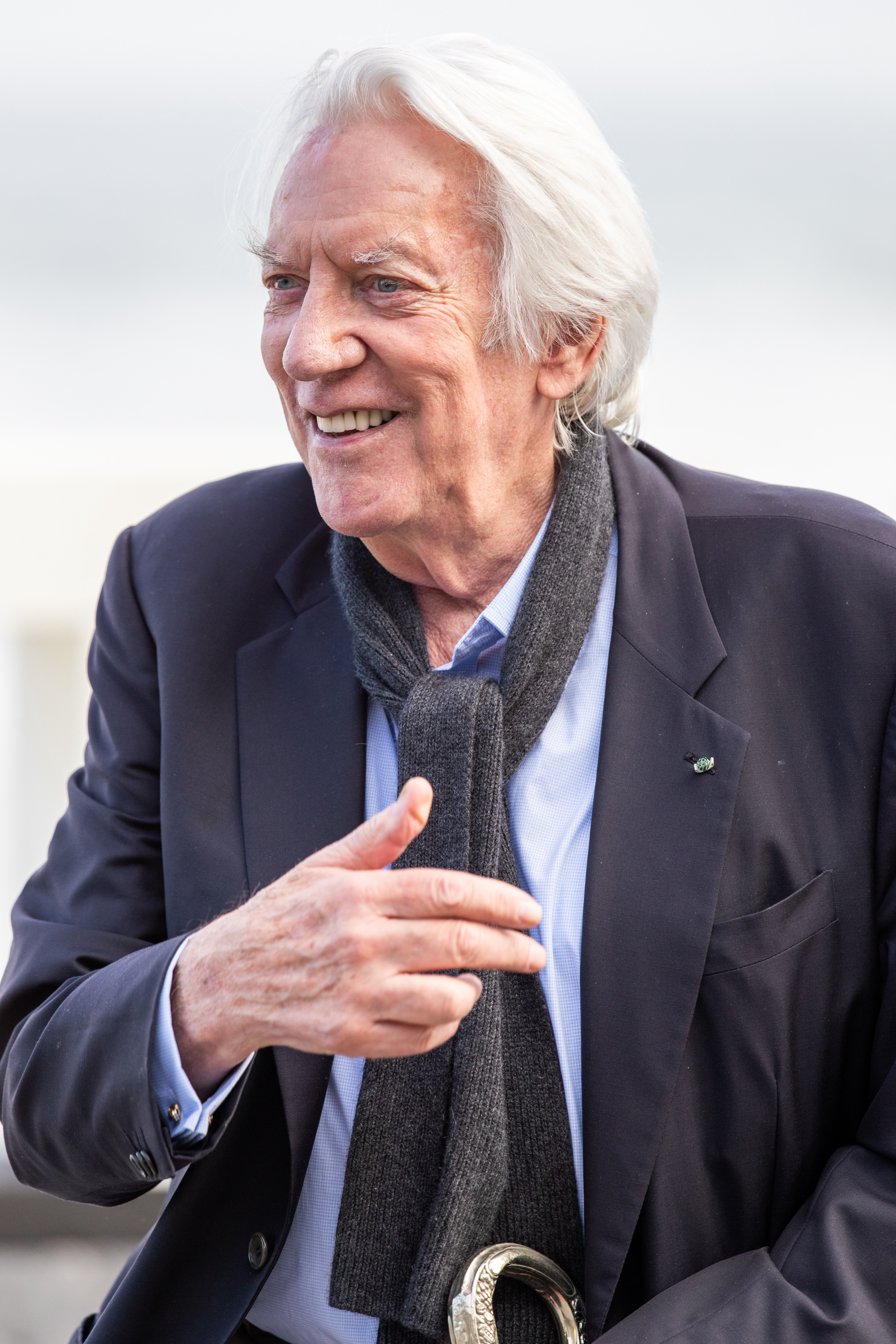 Older man with white hair laughing, wearing a blazer, shirt, and scarf outdoors