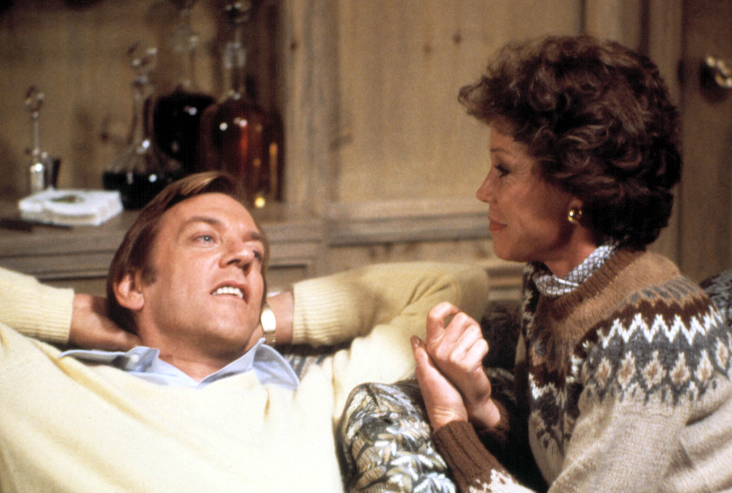A man relaxes on a couch while a woman sits beside him, holding his hand. Both are engaged in conversation