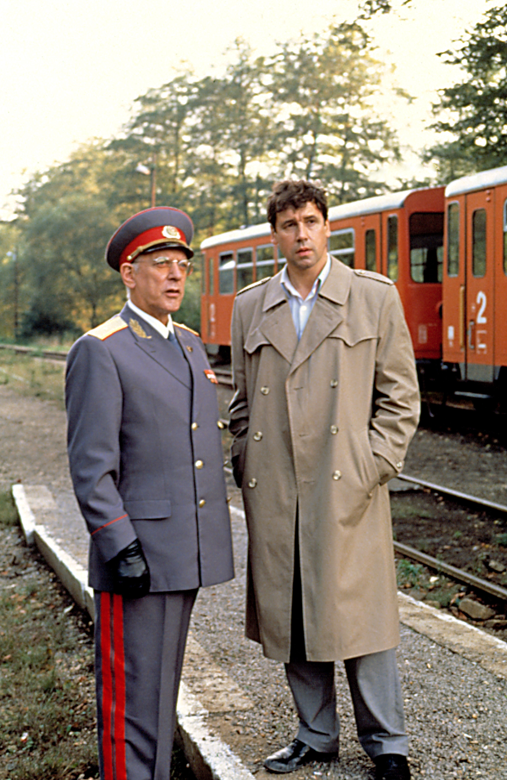 Two men standing on a train platform, one in a military uniform with a peaked cap, the other in a trench coat
