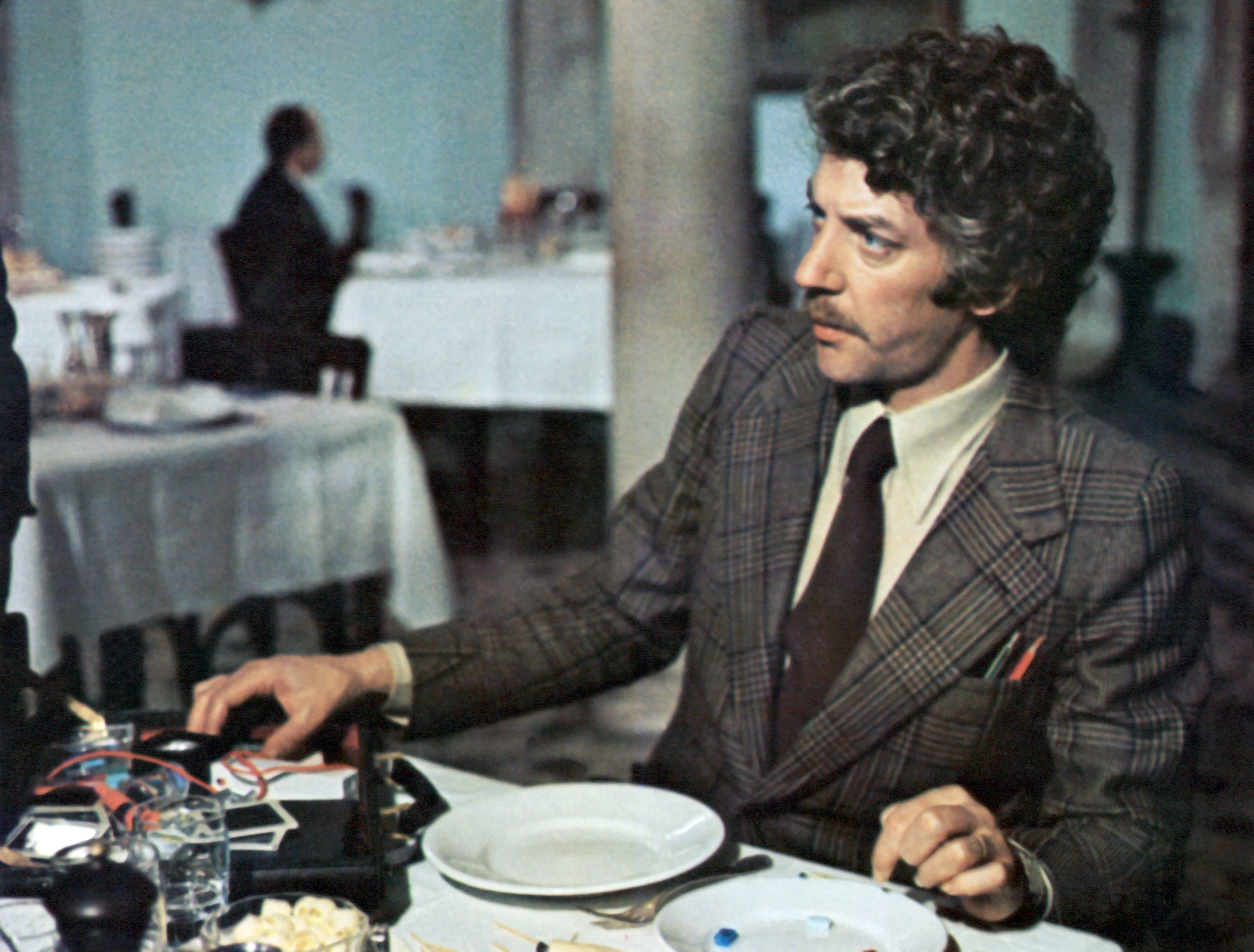 Donald Sutherland sits at a dining table in a formal setting, wearing a plaid suit and tie, with dining utensils and dishes in front of him