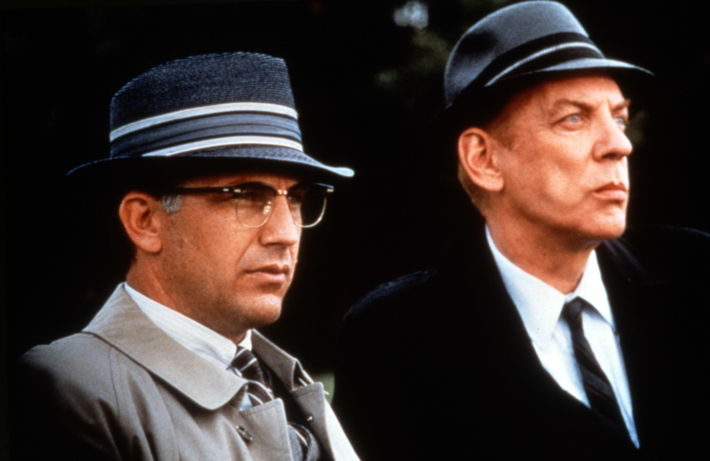 Kevin Costner and Donald Sutherland wear vintage-style 1940s attire with trench coats and fedora hats in a scene from a film