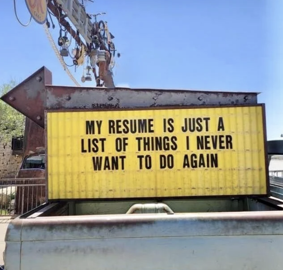 A large sign reads, "My resume is just a list of things I never want to do again." The sign is mounted on a structure near some industrial equipment