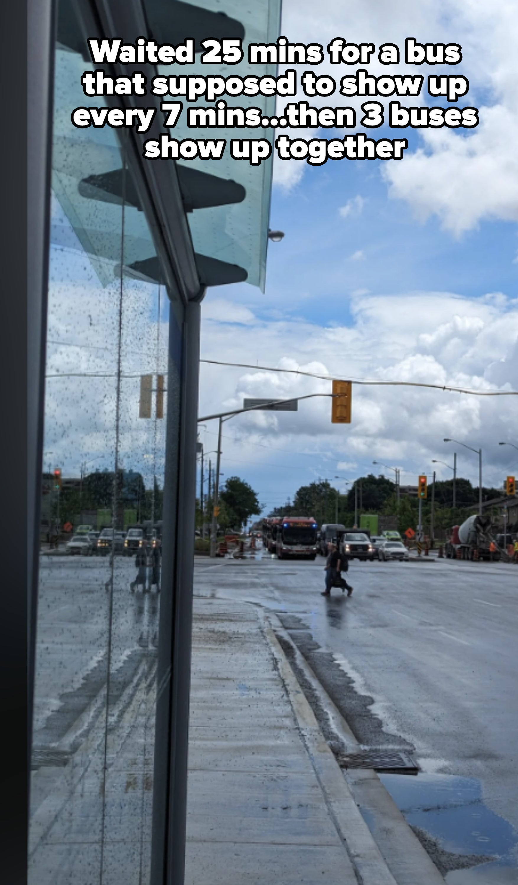 A rainy city street with traffic lights and vehicles in the distance. A person walks on the wet sidewalk near a glass building