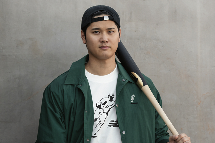 Person in a casual outfit holding a baseball bat on their shoulder. They wear a cap, jacket, graphic tee, and pants