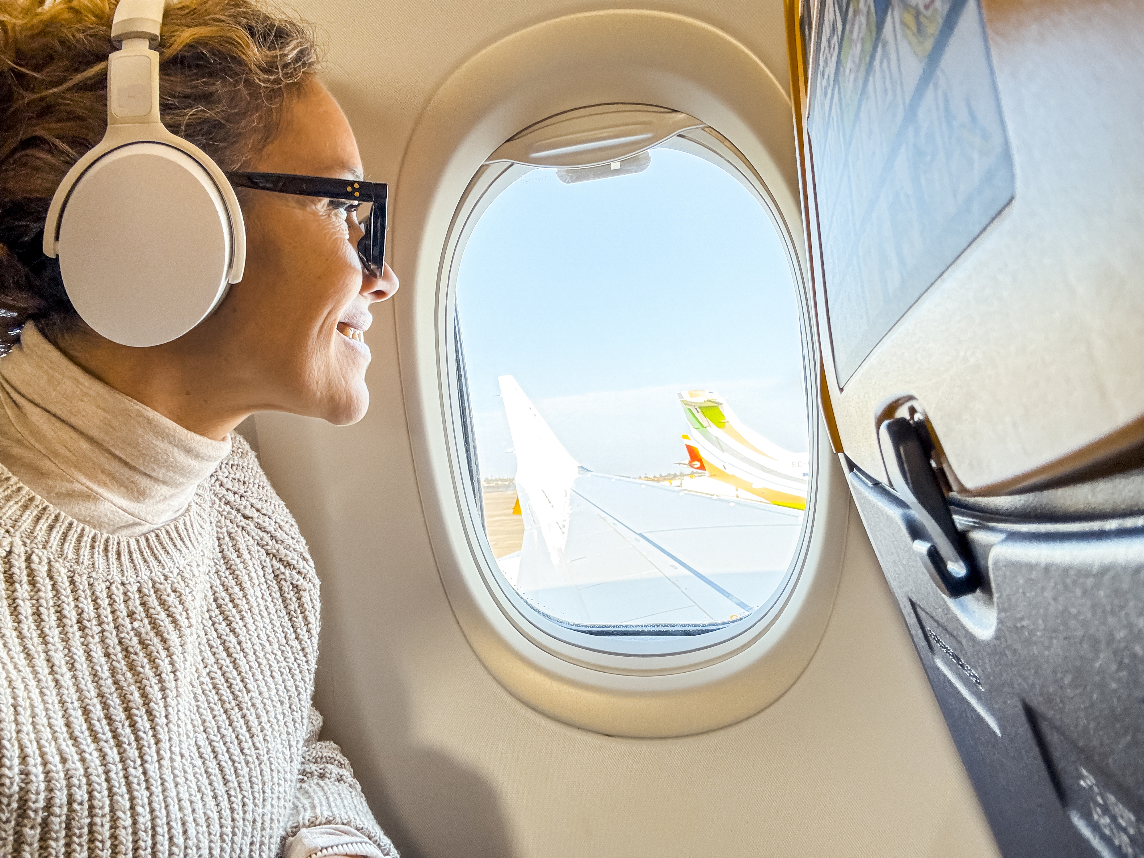 Woman on a plane wearing headphones and sunglasses, looking out the window at an airplane wing