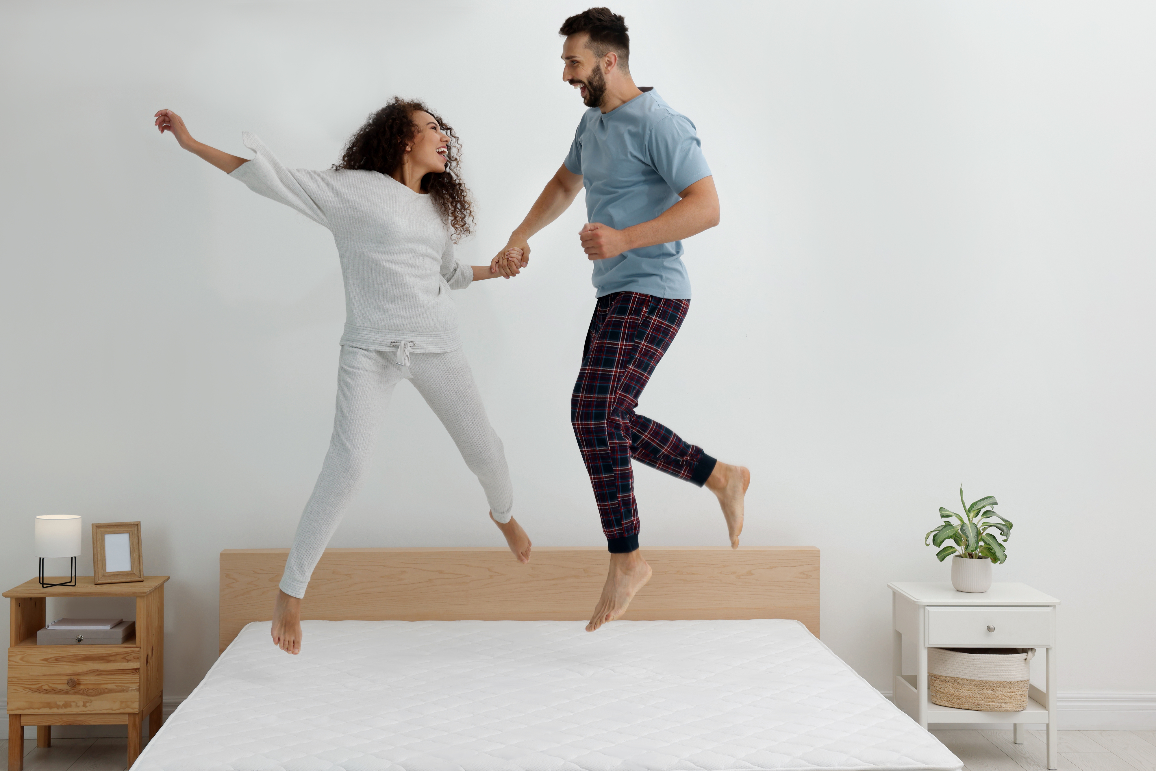 A man and woman happily jump on a bed while holding hands, dressed in casual loungewear. A nightstand and a plant are in the room