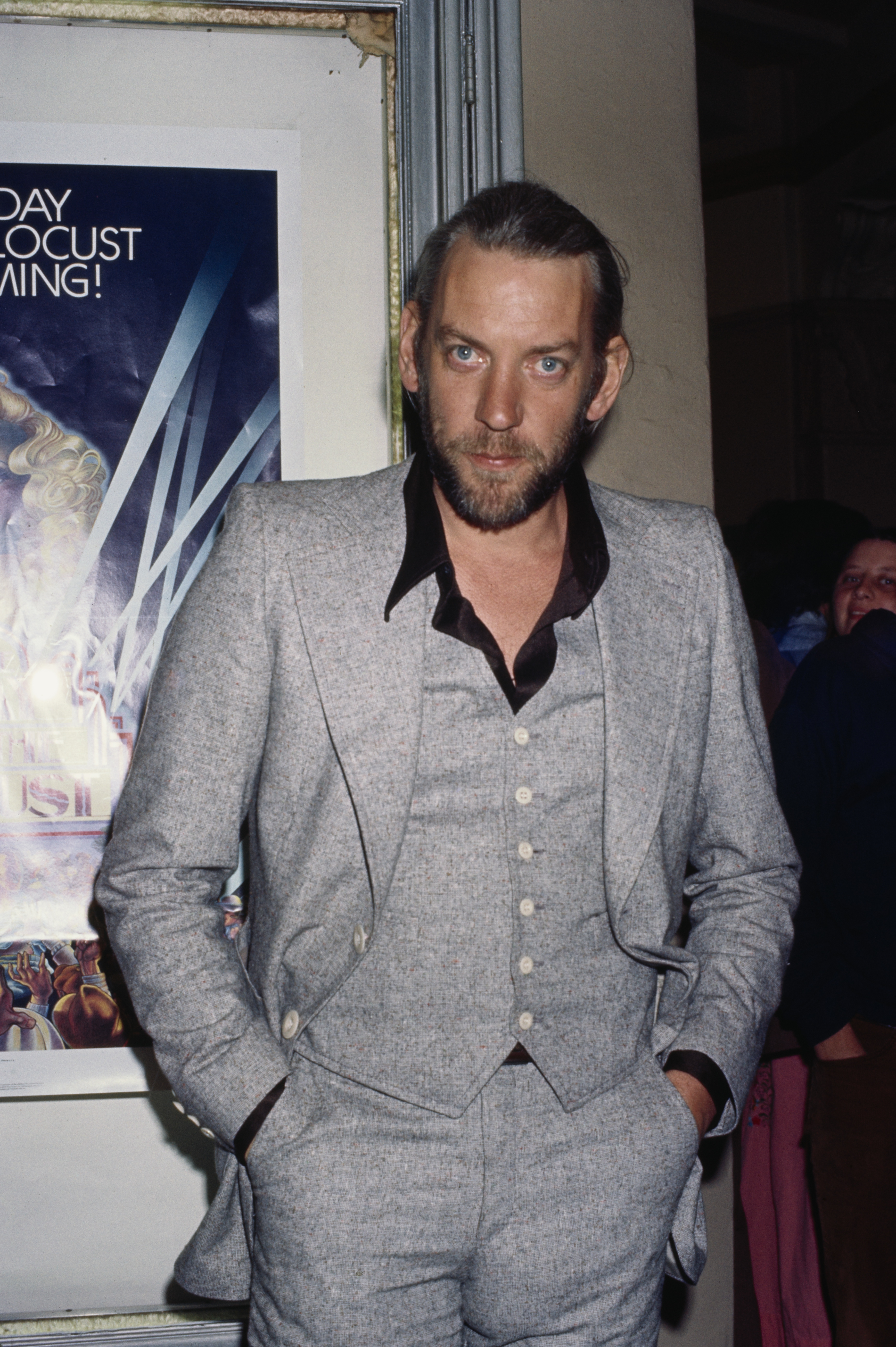 Donald Sutherland poses in a three-piece suit with hands in pockets, standing next to a framed poster