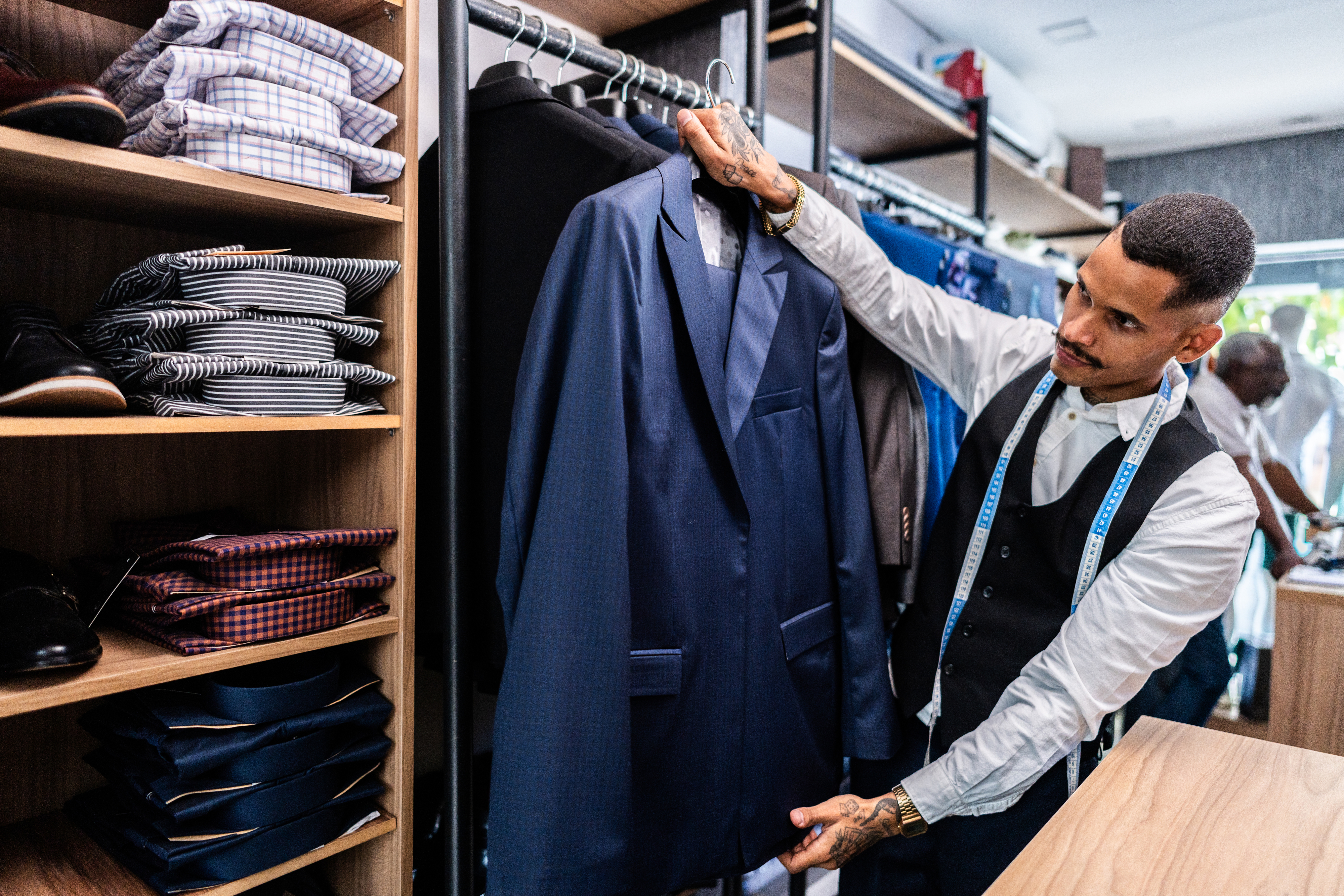 A man in a tailor shop, examining a suit jacket, with shelves of folded shirts and fabrics around him. He wears a vest over a shirt with a tape measure around his neck
