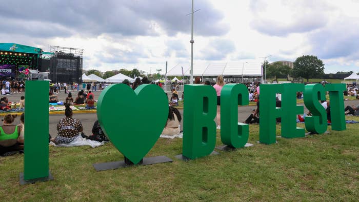 Large green letters in a grassy area spell &quot;I ♥ BC FEST&quot; with a festival stage and attendees in the background at an outdoor event