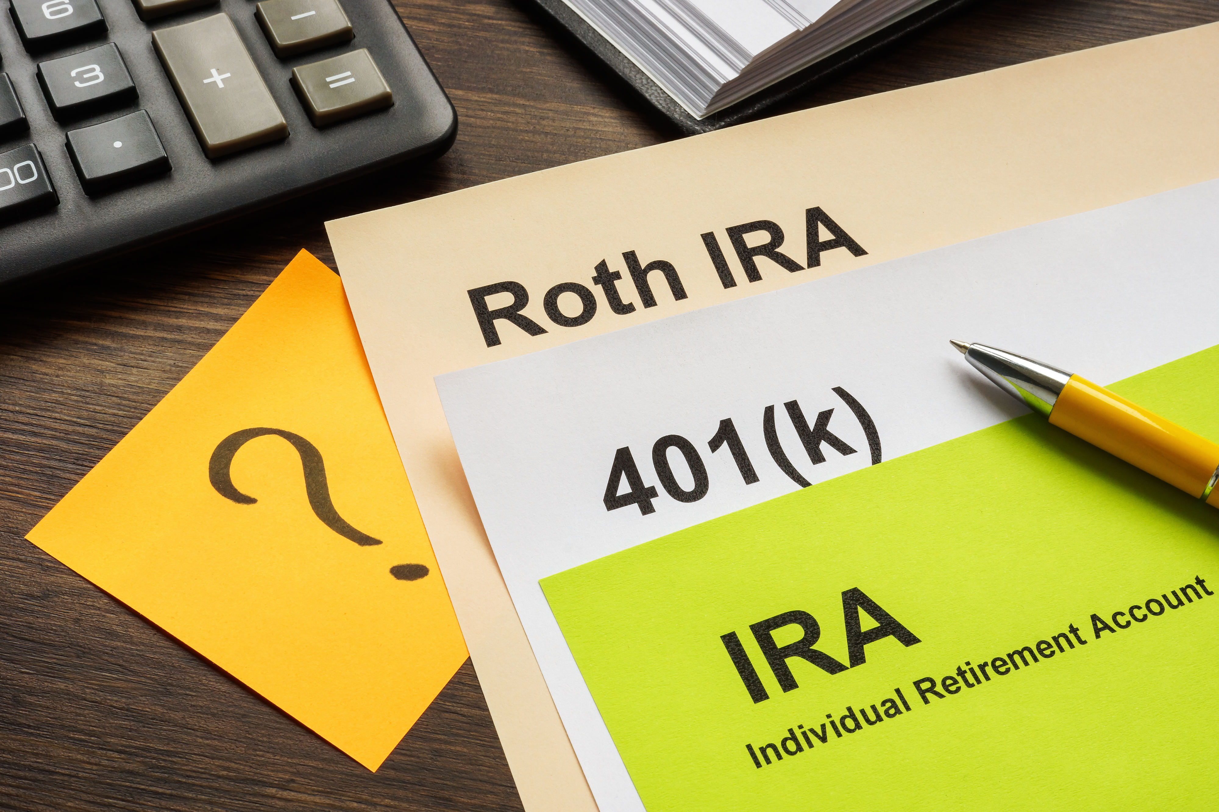 Desk with papers labeled "Roth IRA," "401(k)," and "IRA," a pen, a sticky note with a question mark, part of a calculator, and a notebook
