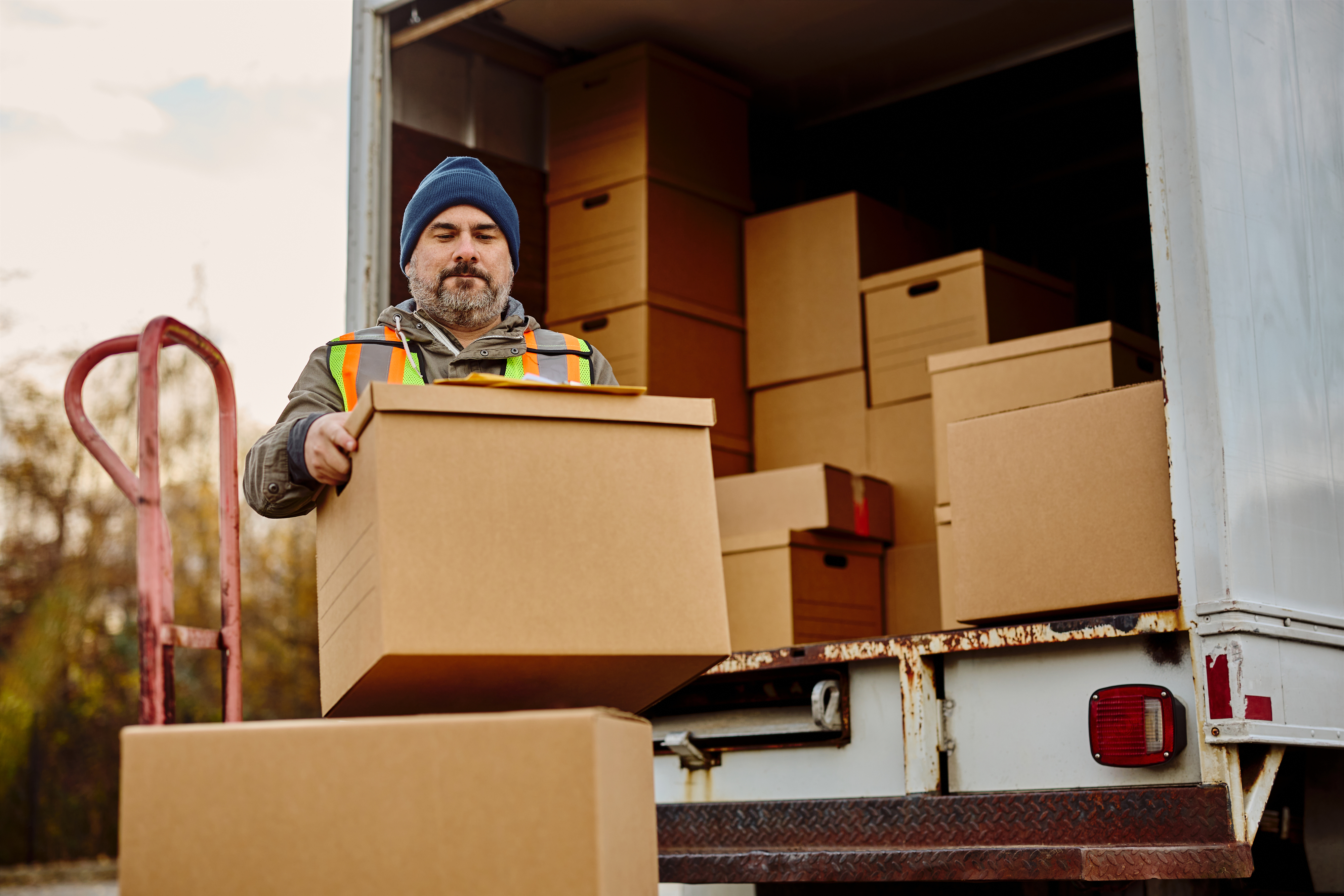 A worker wearing a beanie and a high-visibility vest is unloading boxes from a delivery truck. The image is related to the field of logistics and labor