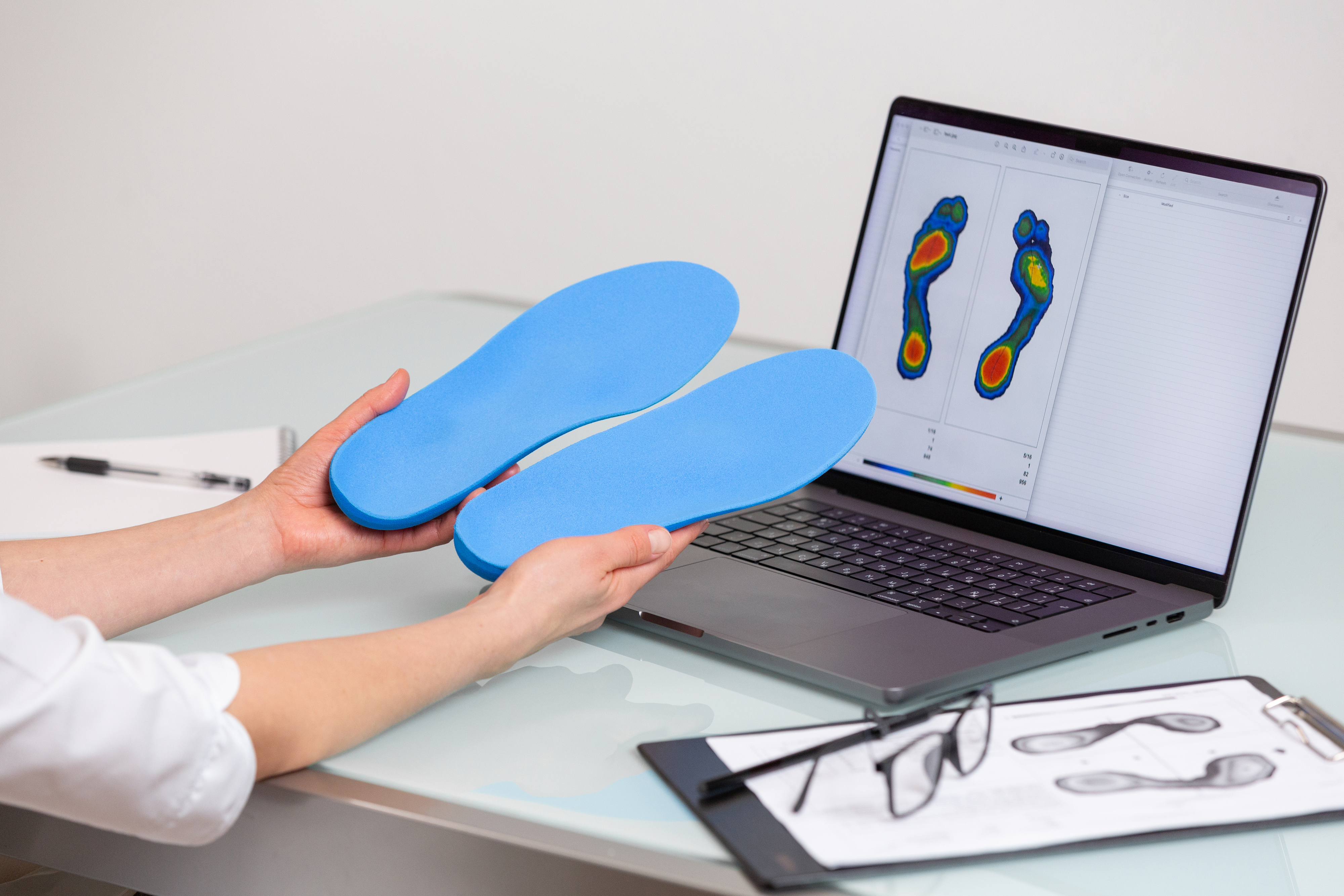 A person holds blue orthopedic insoles in front of a laptop displaying a foot pressure analysis chart. Nearby are a clipboard with a pen and glasses