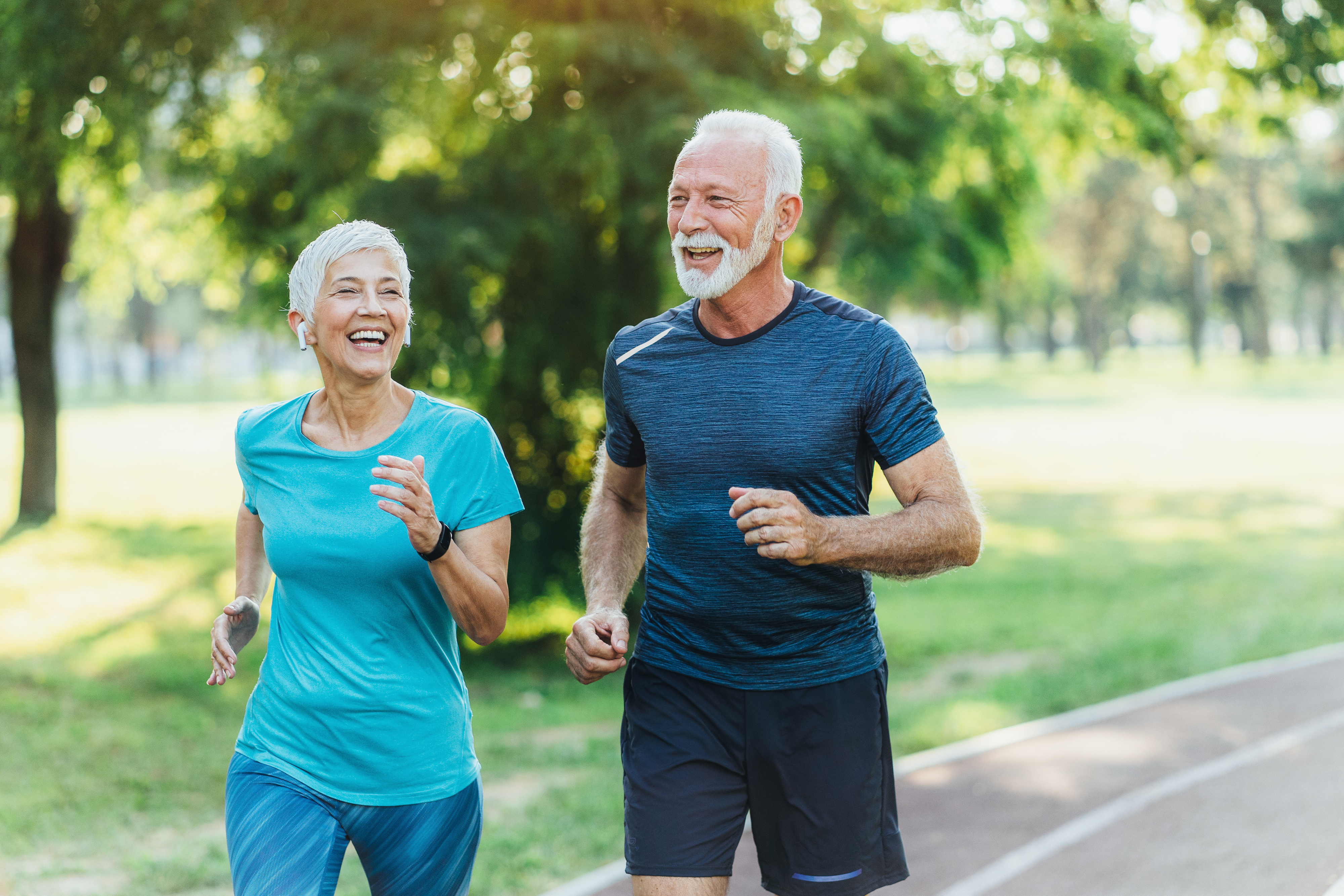 Two elderly individuals, a woman and a man, jog together outside on a park path, smiling and engaged in conversation