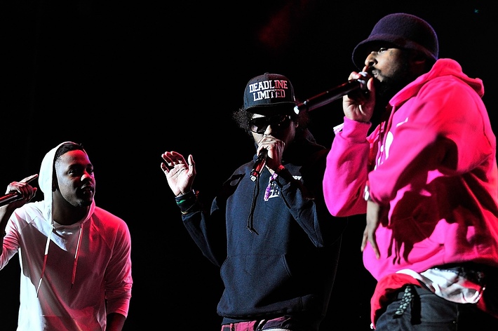 Kendrick Lamar, Ab-Soul, and Schoolboy Q perform on stage. Kendrick Lamar wears a hoodie, Ab-Soul wears sunglasses and a cap, and Schoolboy Q wears a beanie