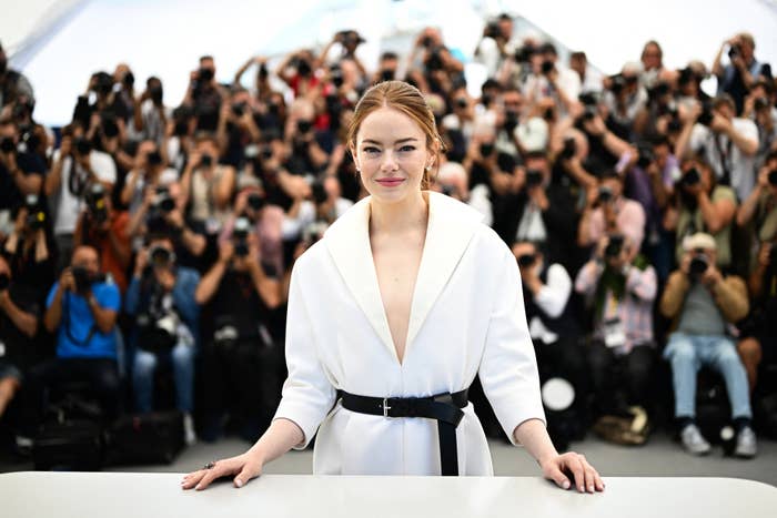 Emma Stone, in a belted outfit, poses at a photo call with a crowd of photographers in the background