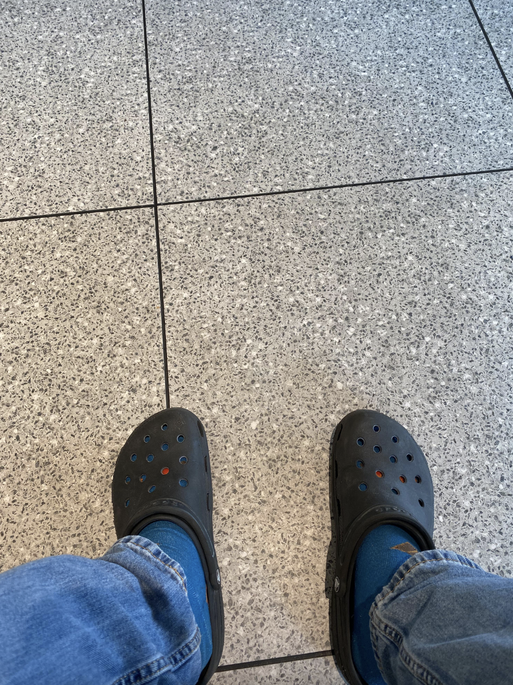 Someone wearing blue socks, grey Crocs, and blue jeans is sitting on a tiled floor