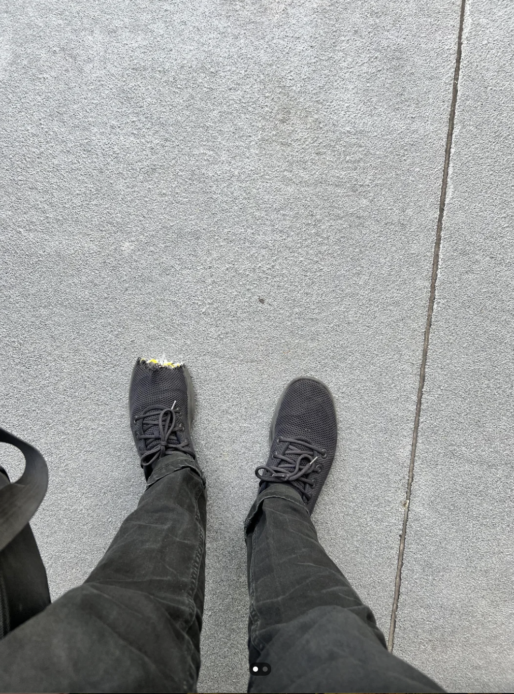 A person's legs and sneakers are seen from above. The left shoe is damaged at the toe, revealing part of a sock. The sidewalk is visible in the background