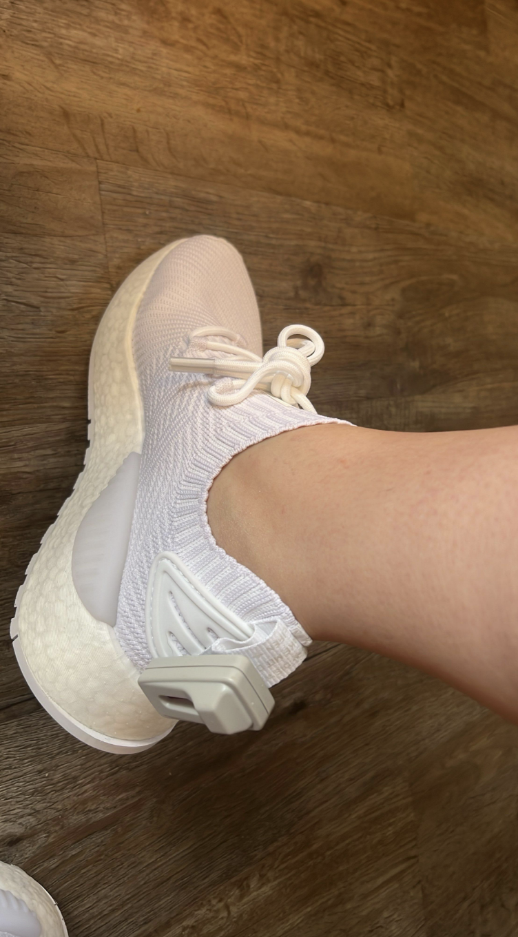 Close-up of a person's foot wearing a white knit sneaker with a high ankle collar, lace-up front, and a thick cushioned sole, on a wooden surface