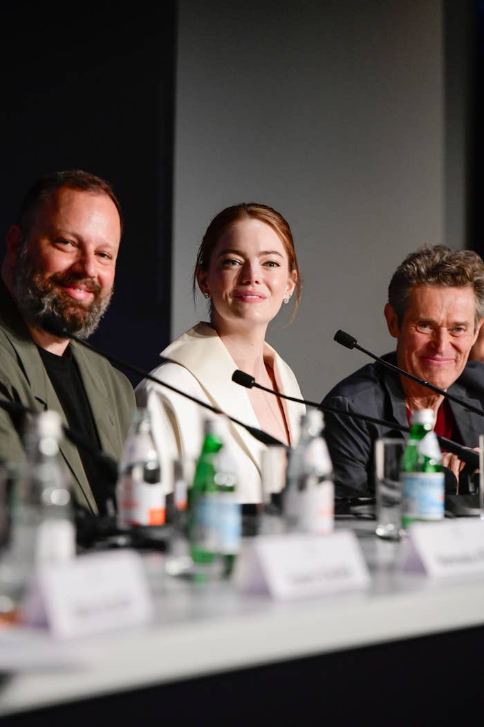 Yorgos Lanthimos, Emma Stone, and Willem Dafoe at a press conference, smiling and seated behind microphones and water bottles