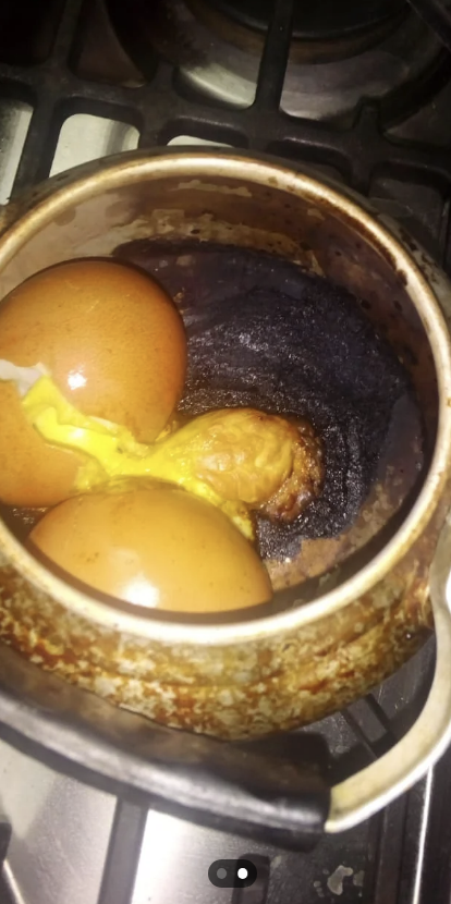 Two cracked eggs in a burnt pot on a stove with a yolk oozed out