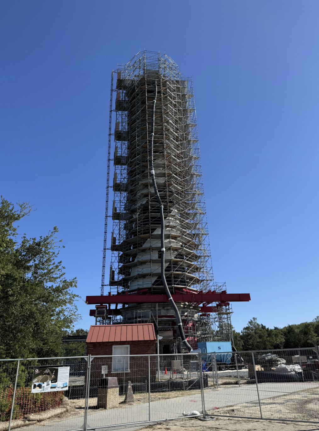 View of a cylindrical structure undergoing maintenance, surrounded by scaffolding and fencing with visible signage. Trees and a clear sky are in the background