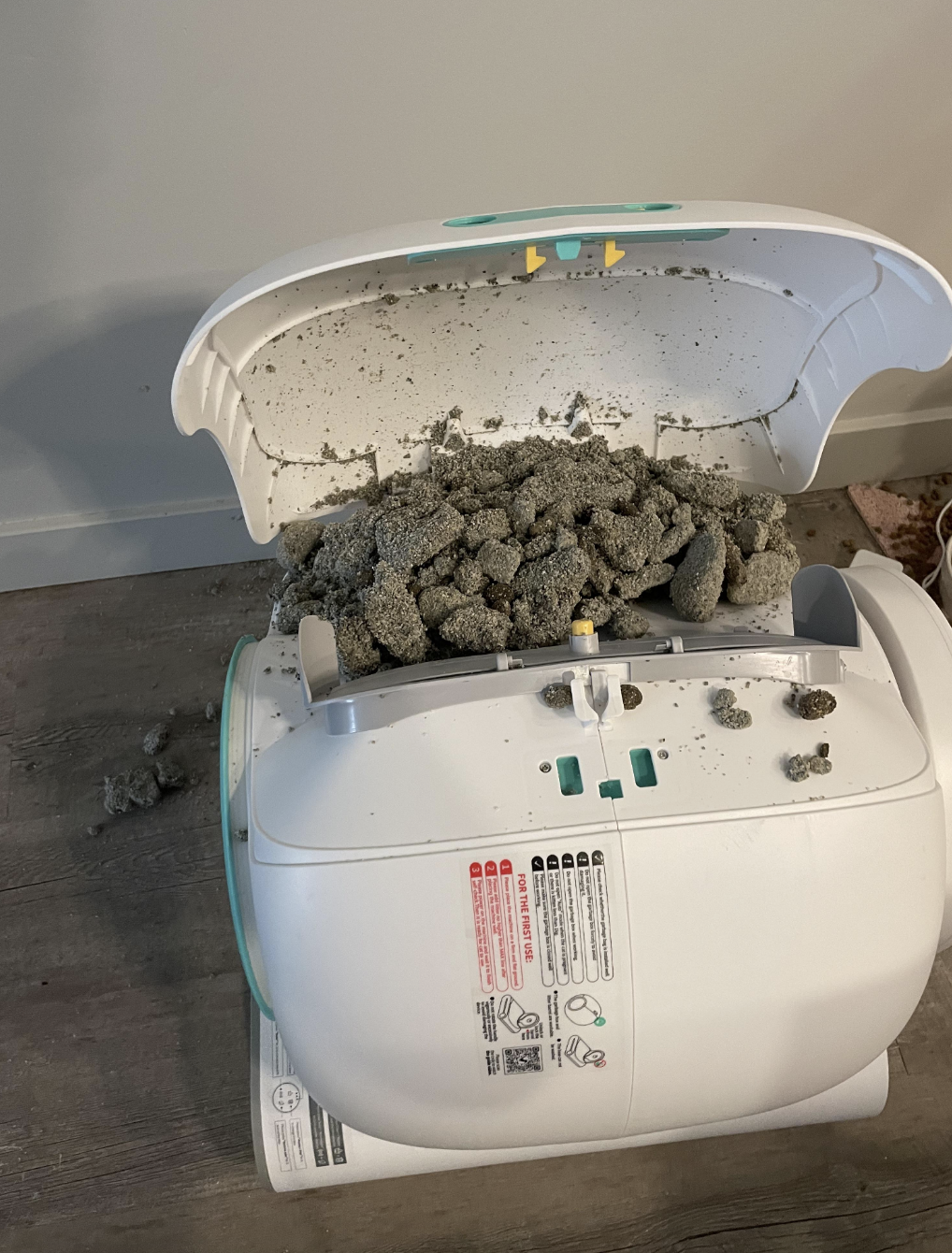 Broken automatic cat litter box with clumped litter overflowing onto the floor