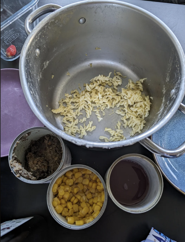 A cooking pot with some cooked pasta, surrounded by open cans containing corn, ground meat, and a dark sauce. A strawberry is visible in the background