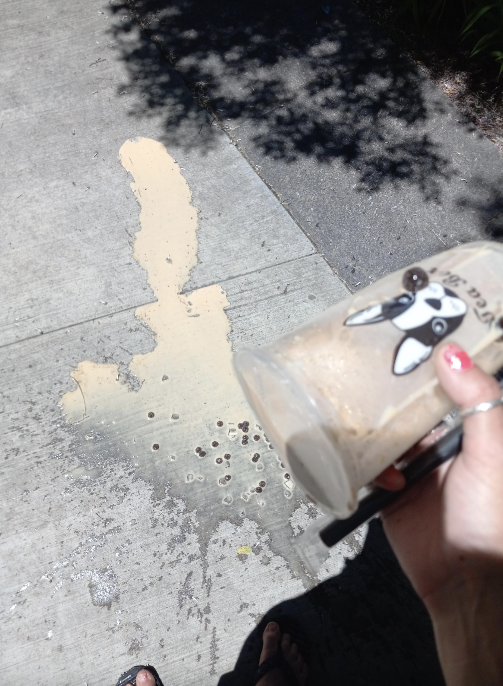 Person accidentally drops a partially consumed boba tea on a sidewalk, with the spilled drink spread across the concrete. The person's hand is visible, holding keys