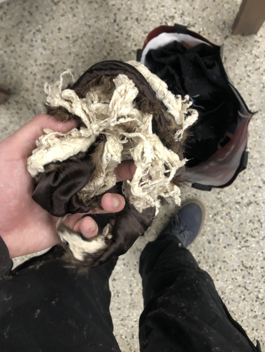 A hand holding a tangled, worn mop above a basket. A person in dark clothing and sneakers is visible from the waist down